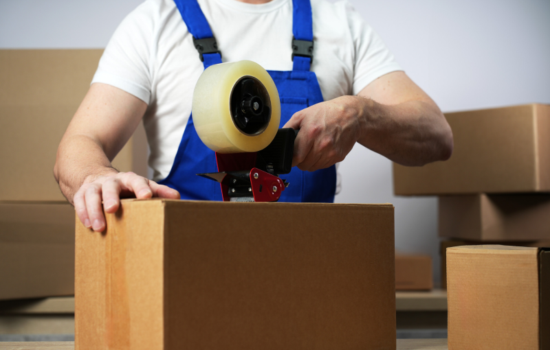 Worker sealing box with tape for moving, close-up. Hands of postal male worker in uniform packing carton box at delivery department in post office. Online shopping and parcel shipping concept